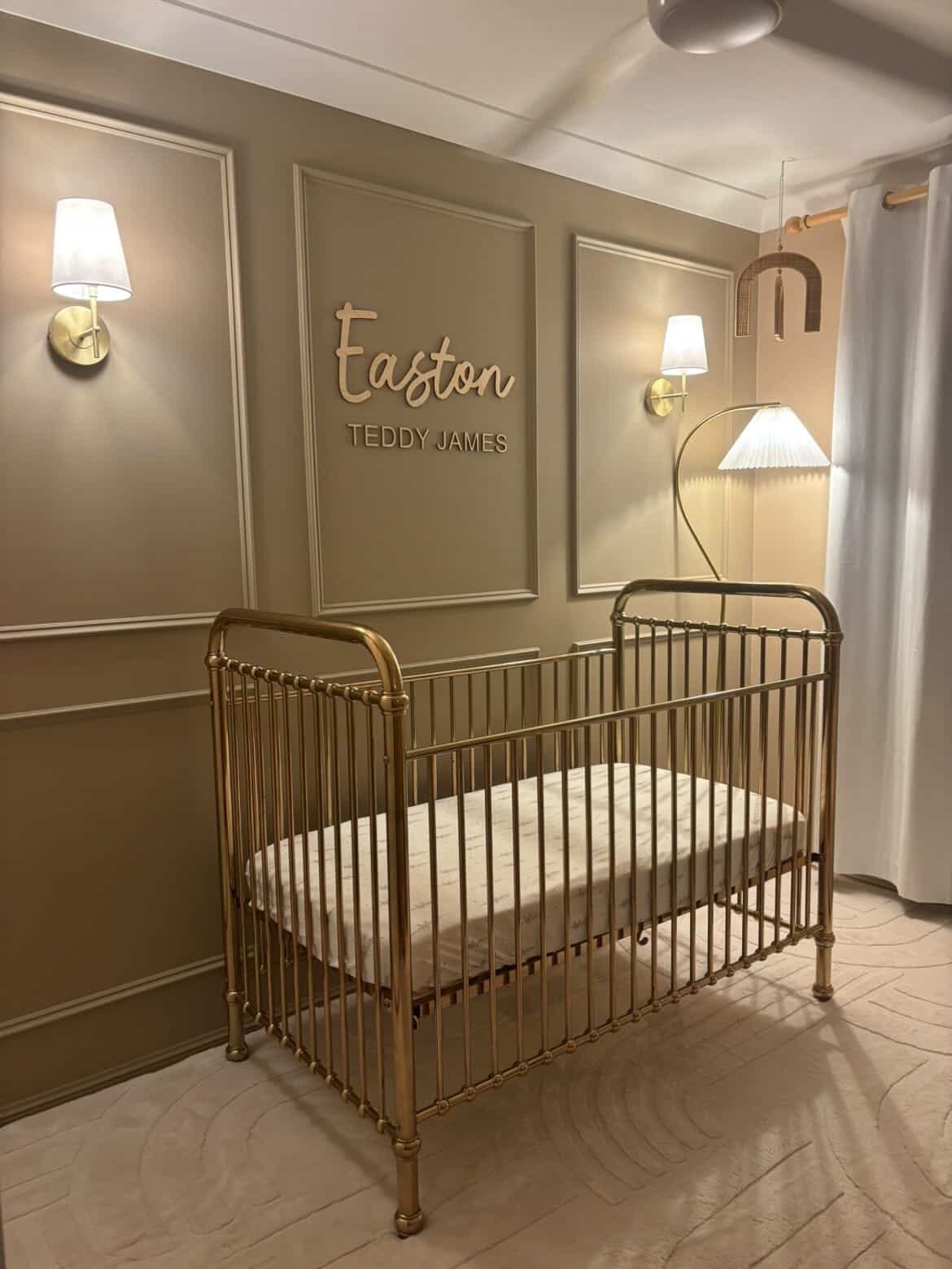A gold metal crib with a white mattress in a styled nursery featuring beige walls, decorative molding, wall sconces, and a custom name sign reading 'Easton TEDDY JAMES'