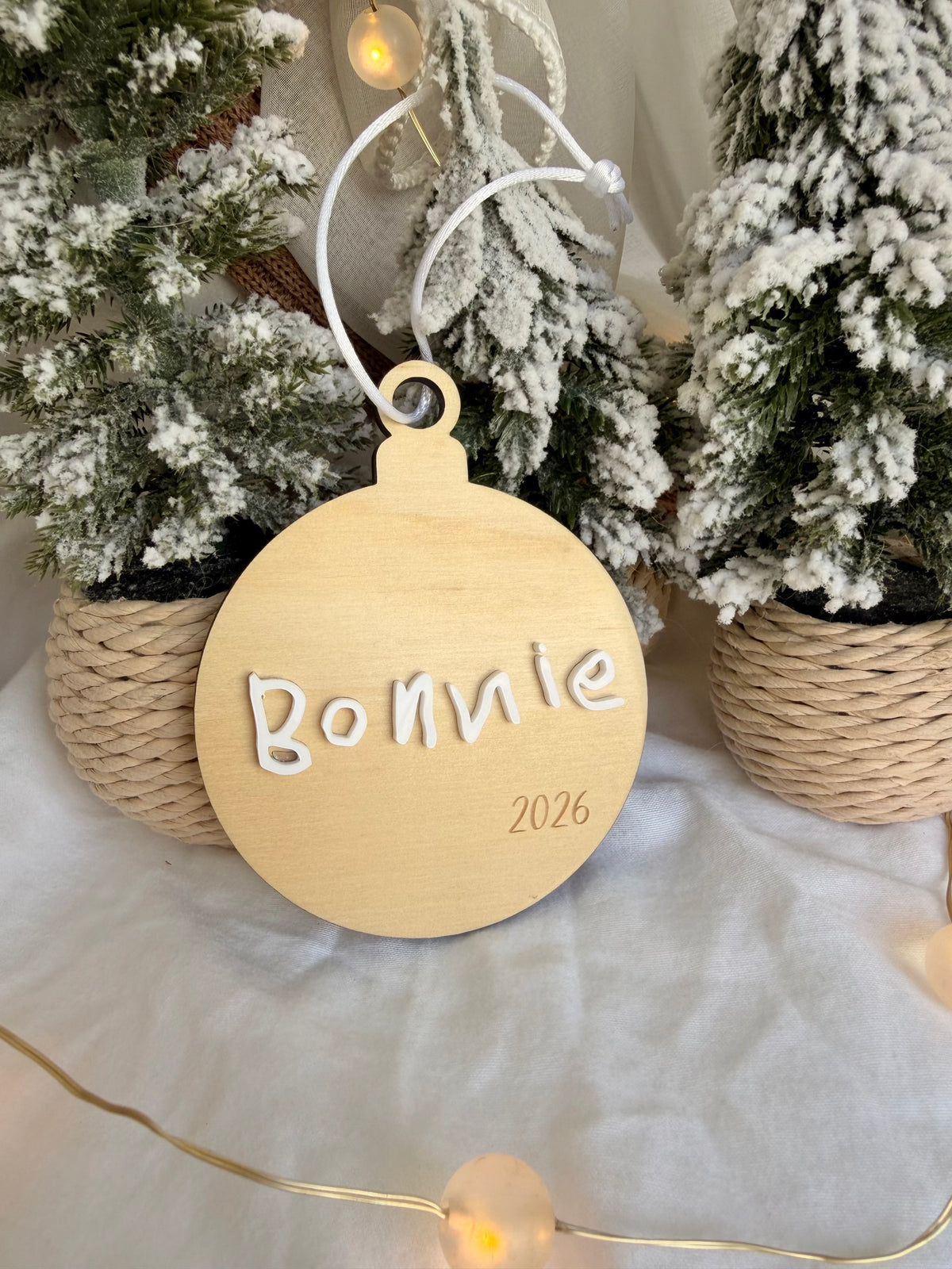 A personalized round wooden ornament hanging in front of frosted Christmas trees. The ornament is light natural wood with the name 'Bonnie' and the year '2026' engraved in white, suspended by a white string.