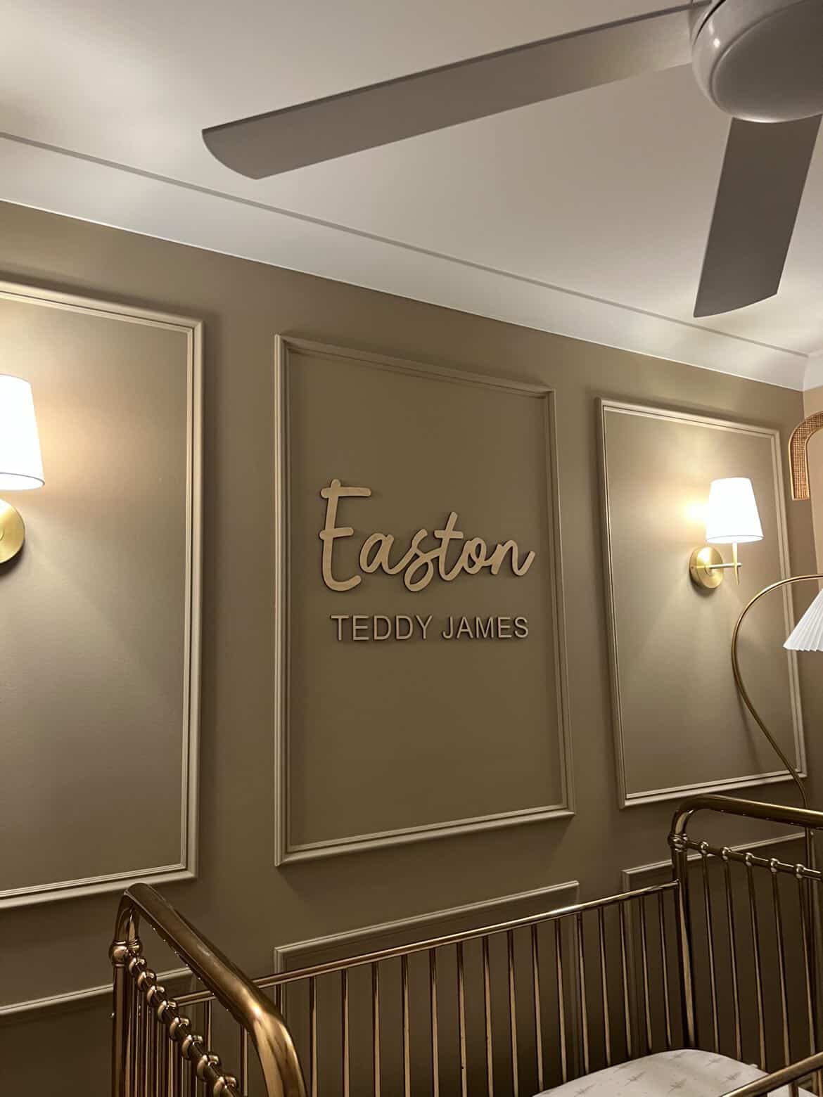 Custom wooden wall sign in a nursery setting, featuring the name 'Easton' in a cursive font and 'TEDDY JAMES' in a serif font, mounted on a beige paneled wall above a gold crib.