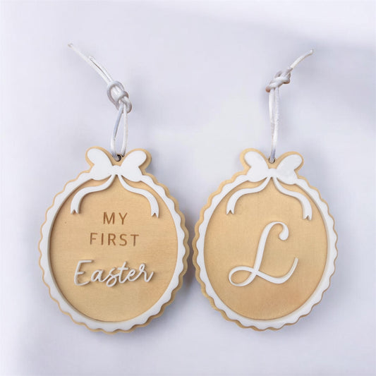 Flat lay of two double sided personalised timber easter ornaments with white acrylic bow detail and scalloped edges, featuring "My First Easter" on one side and a custom initial on the reverse, hanging with white ribbon