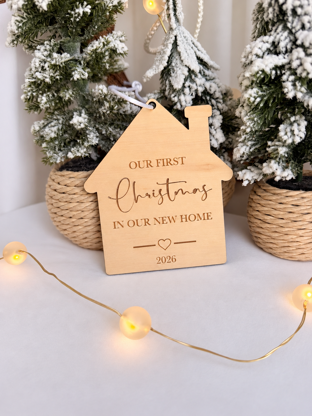 Wooden house-shaped Christmas ornament engraved Our First Christmas in Our New Home 2026, displayed with snowy Christmas trees, handmade by Ally May Creations Australia.
