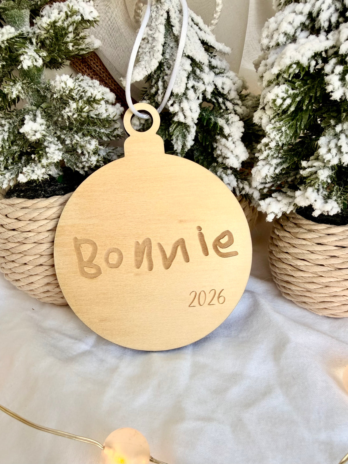 A personalized round wooden ornament engraved with 'Bonnie 2026' and hanging from a white string, styled with frosted miniature trees and a white fabric backdrop.